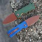 SHOOZIZ HAN306-GMRG and LMRG folding knives open on stone, 14C28N steel with rose gold PVD, green and blue Micarta handles.