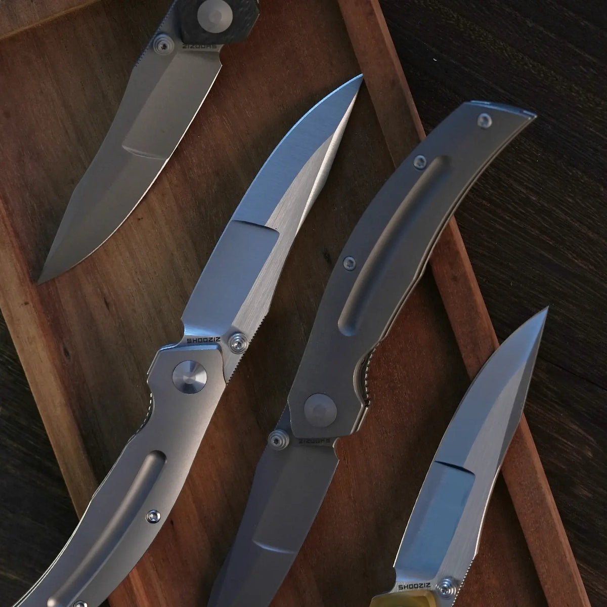 SHOOZIZ SERPENT series folding knives on wooden board, featuring TC4 titanium and yellow carbon fiber handles with satin ELMAX steel blades.